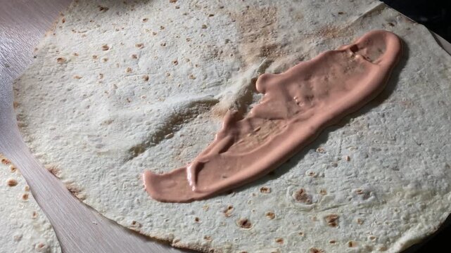 Close-up of a spoon spreading pink creamy sauce over a round wheat flatbread. Preparation of homemade shawarma, wrap, or burrito on a table
