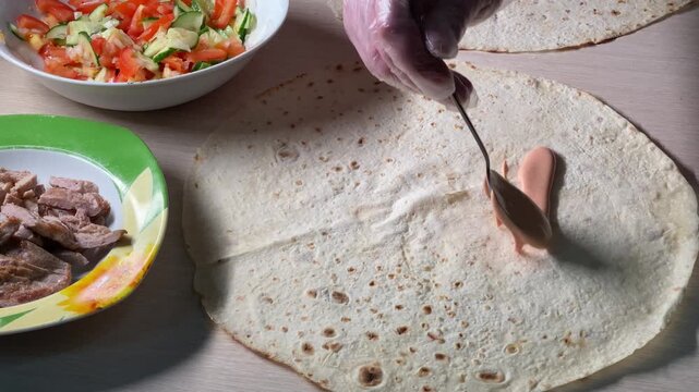 Spreading pink sauce on a flatbread with a spoon. Making homemade shawarma or tortilla wrap with ingredients like meat and salad in the background.