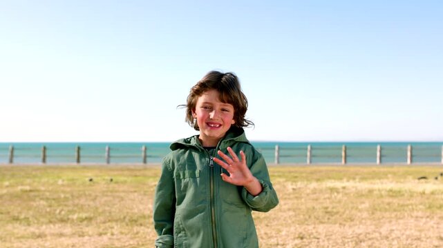Outdoor, face and boy with wave for greeting, travel destination and winter holiday for weekend trip. Beach, space and happy child with hello gesture for video call, nature and portrait for vacation