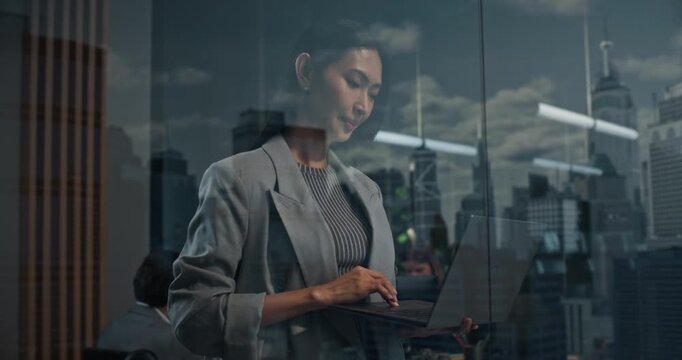 Asian Businesswoman Stands With a Laptop by an Office Window, Thinking Through Startup Growth Strategy as Her Leadership Focus Brings Success. Cinematic Reflections of Big City Financial District