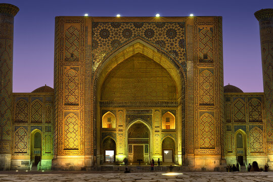 Majestic illuminated Ulugbek Madrasah facade on Registan Square at evening with blue pink sky. Golden ornate arch, minarets, blue mosaic tiles. Islamic heritage, landmark of Samarkand, Uzbekistan.