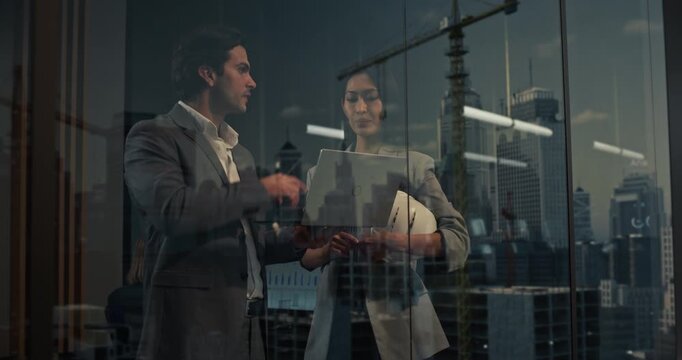 Caucasian Businessman and Asian Businesswoman Analyze a Laptop by a Tower Window, Planning Finance and Startup Growth While City Glass Reflects Cranes, Offices, Investors and Urban Success.