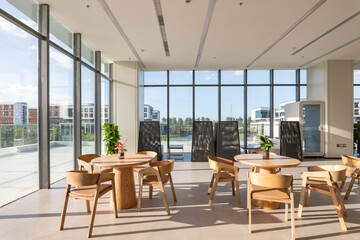 Fototapeta premium Modern office lounge with wooden round tables and chairs bathed in natural sunlight through floor-to-ceiling windows