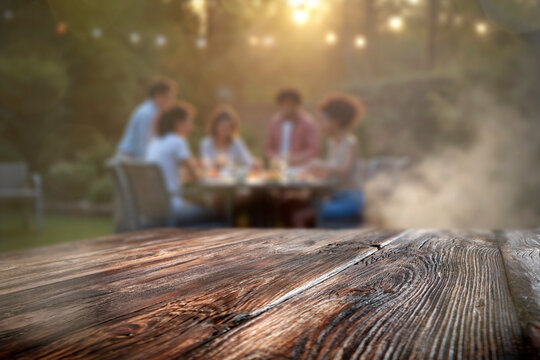Empty wooden table with clear copy space for product placement, set outdoors during spring or summer. Blurred background of barbecue scene suggests grilling, outdoor dining and relaxation.