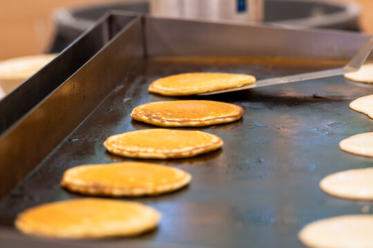 A neat row of pancakes fills a flat top grill, and at the far end a long spatula is mid-flip, catching the furthest pancake in the moment of rotation. 