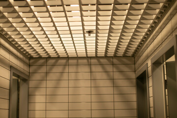 Obraz premium Interiors. Textured ceiling grid casts dramatic shadows in corporate corridor with tiled walls, warm sepia tones, soft ambient lighting and quiet minimalist