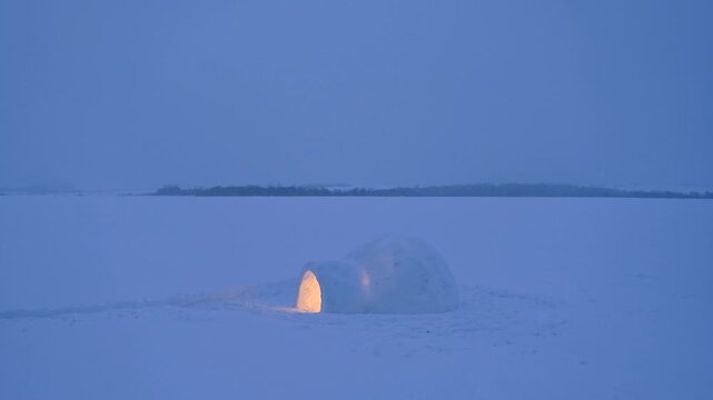 Lonely igloo glowing with warm light on frozen snowy field during snowfall. Winter night landscape, snow falling over arctic style shelter