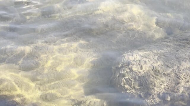 A beautiful close-up abstract shot of gentle water ripples with bright sunlight creating intricate caustic light patterns on the shallow riverbed. The shifting golden light and soft shadows dance acro