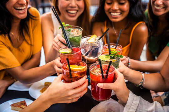 Happy people toasting cocktail glasses sitting at bar table - Cheerful friends enjoying happy hour at pub restaurant balcony - Food and beverage life style concept