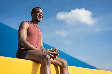 Happy smiling African man sitting on colourful yellow and blue wall after running and doing sport workout