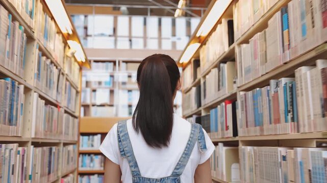 Back view of young student woman walking between the bookshelves, walks down a long library aisle filled with numerous books, looking for the necessary book in modern bookstore interior scene