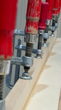 A row of red-handled C-clamps firmly holds wooden planks in place. Metal parts show slight wear. The setup suggests woodworking or joinery work in progress, emphasizing precision and stability.