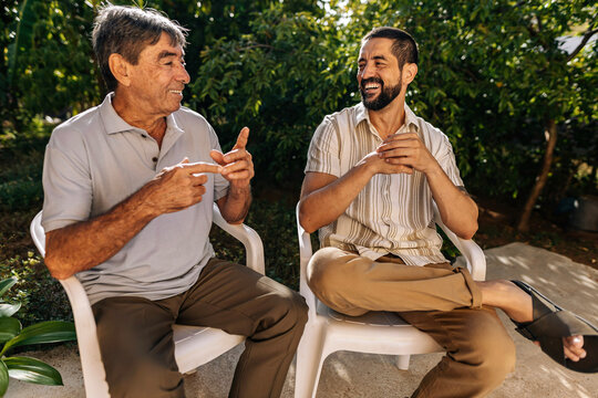Senior father and adult son sitting outdoors talking and laughing together, enjoying a relaxed family moment and connection in a garden setting.