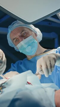 Vertical low angle shot of male surgeon with sterile mask having conversation with his assistant while they preforming operation on patient