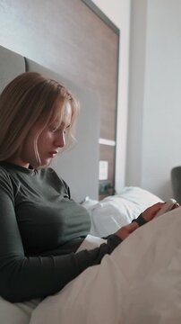 A beautiful young woman lies in bed, chatting online and smiling. A woman with a phone in a hotel room reads messages in the morning.