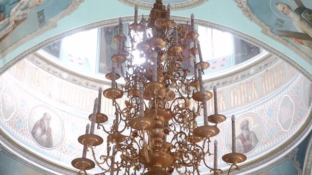 Majestic golden chandelier with multiple tiers and candles hanging from light blue cathedral dome decorated with religious frescoes depicting saints and cyrillic inscriptions