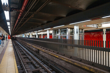 Fototapeta premium The Bowling Green subway station platform in Manhattan shows curved underground train tracks and red tiled walls. Fluorescent lights illuminate the New York City metro transportation system.