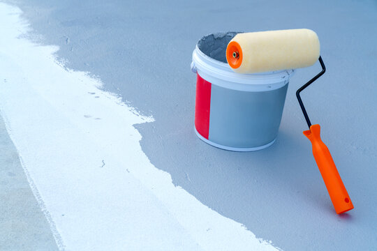 Orange paint roller resting on a bucket of grey liquid coating on a rooftop or floor. A white stripe of primer is visible on the textured surface. Concept of waterproofing and renovation.