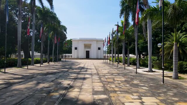 A slow forward walk toward a white mausoleum across a tiled stone plaza flanked by tall palm trees and rows of flagpoles under a clear blue sky.