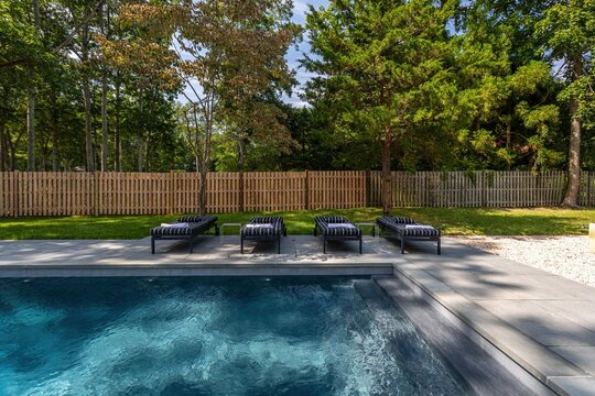 Modern backyard swimming pool with lounge chairs, paved patio and wooded fence line