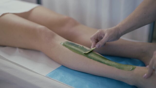 A cosmetologist applies green sugar paste to a woman's leg for hair removal. This professional sugaring service takes place in a clean beauty salon environment.