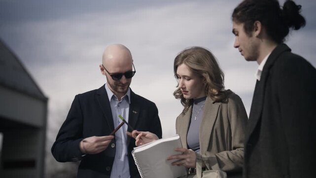 Three business professionals discuss matters during an outdoor meeting. They are collaborating and planning, reviewing documents in a professional setting.