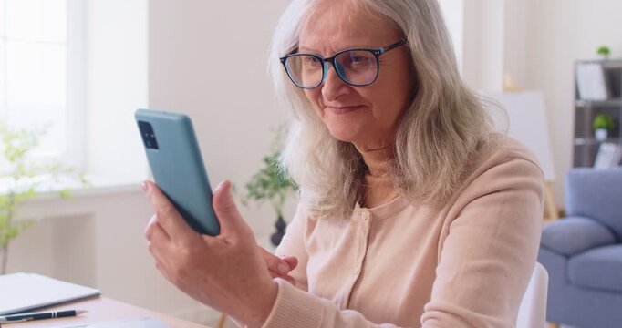 Senior woman with poor vision trying to read text on phone screen. Elderly lady with presbyopia or eyesight problem sitting at home table, wearing glasses and holding smartphone farther to focus