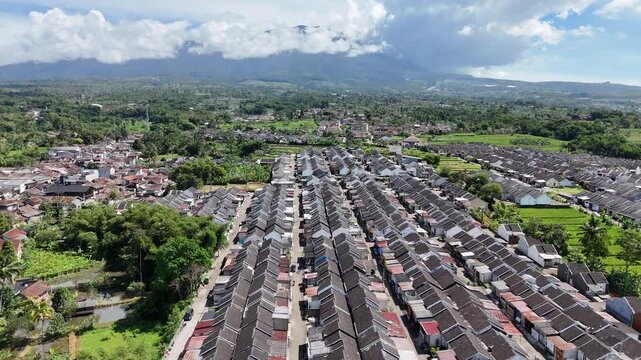 An impressive aerial perspective showcases rows of symmetrical suburban houses leading the eye toward a massive, cloud-shrouded mountain peak under a bright, clear blue sky in a tropical landscape.