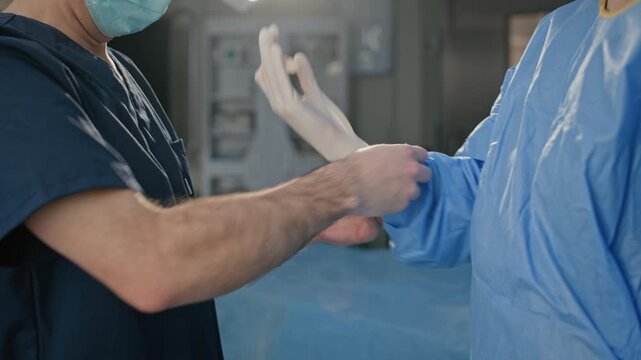 Caucasian assistant in scrubs helping female surgeon pull on sterile latex gloves before operation and having talk