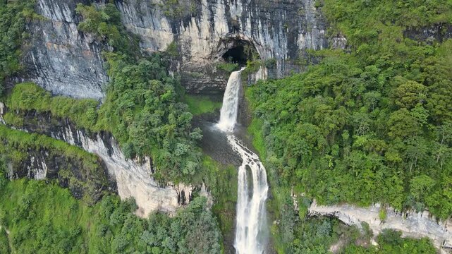 Drone footage of Tisquizoque waterfall pouring from a cave in sheer limestone cliffs, surrounded by dense green forest and a dramatic two-tier cascade.