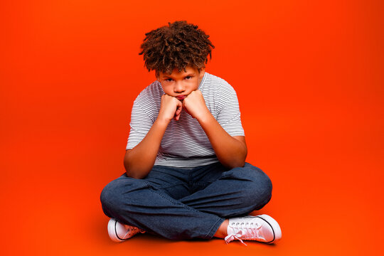 Pensive boy sitting cross-legged on an orange background looking bored and upset in casual striped shirt, jeans and white sneakers