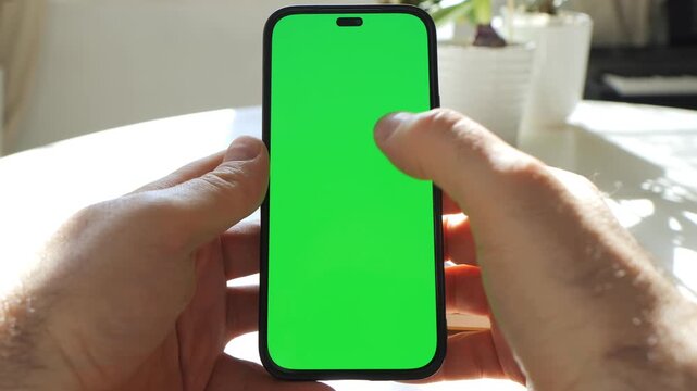 Hands hold smartphone with green screen. Phone held steady, then finger taps screen. Background shows table and potted plant. Natural light illuminates scene