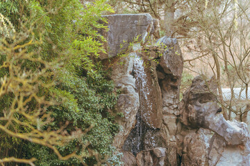 Water cascades down a stone structure. Lush green foliage surrounds the formation. Sparse branches contrast with the vibrant plants. This tranquil natural scene invites calm reflection © Jakob
