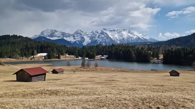 wide-angle landscape of the scenic Geroldsee lake in the Bavarian Alps near Garmisch-Partenkirchen. In the foreground, traditional wooden hay huts (Stadel) sit on a rolling golden meadow.