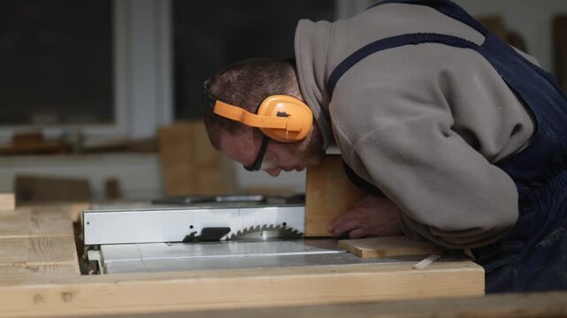 A man meticulously cuts wood with a table saw in his workshop. He wears protective earmuffs and safety glasses while working.