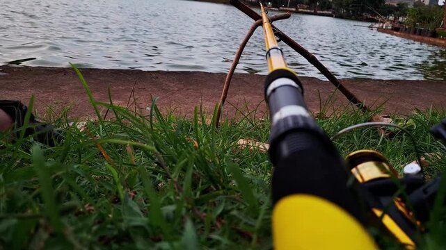 An angler places a fishing rod by the edge of a lake while waiting for fish to take the bait. Fishing activity in calm lake waters with a natural atmosphere, focused on relaxation in nature.
