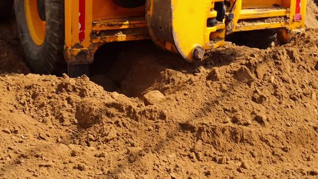 Heavy duty yellow JCB excavator bucket lifting and moving fresh brown soil to level the ground at a residential construction site or plot development project.
