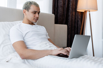 Young man using laptop in bed, working remotely from home in morning