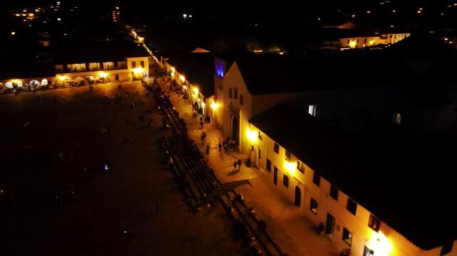 Aerial night view of Villa de Leyva main plaza showing an illuminated church facade and rows of benches under warm streetlights, with small groups of people walking across the square.