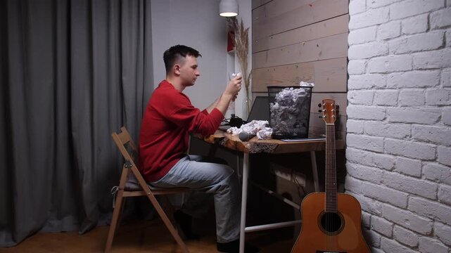 Man at desk crumpling paper in frustration, surrounded by discarded notes and a guitar, struggling with writers block and creative exhaustion while trying to compose music or lyrics