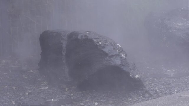 Heavy fog surrounds a large rock. Water droplets glisten on its surface. Damp ground shows pebbles and leaves. The rock's texture appears rough. Fog creates a mysterious atmosphere