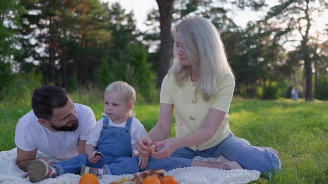 White family picnic with toddler outdoors, mother blonde and father bearded sitting on blanket, child in denim reaching for snack, shared laughter, soft evening light through trees,