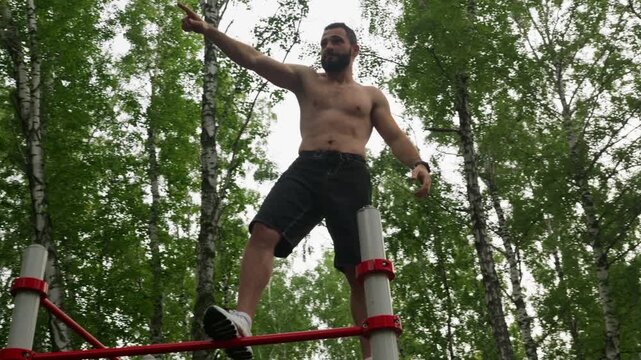 Shirtless male celebrating on top of red exercise frame, arms raised in triumphant pose, smiling against canopy of birch trees, playful acrobat vibe, crowdfree forest setting, energized