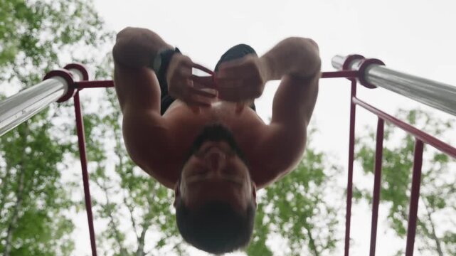 Shirtless white athlete on park bars performing upsidedown hang and core strength routine on red parallel bars beneath leafy canopy, focused expression, sweat on defined biceps, outdoor