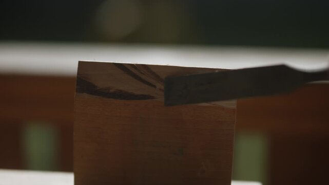 A hand uses a chisel to scrape a small piece of wood, revealing its natural texture. This close-up shot captures the detailed work of woodworking and crafting.