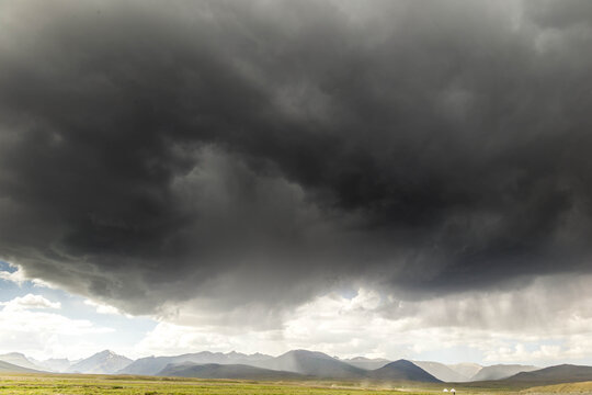 View of an approaching storm hangs heavy above the open green fields and distant mountains, casting shadows across the landscape, Deosai National Park, Pakistan.