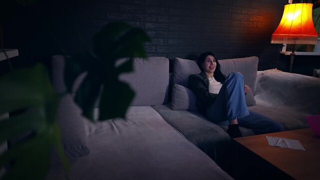 Young adult Caucasian lady is watching a film in the evening at home resting alone in the living room. Pretty woman is spending weekend in front of television and looking surprised by the show