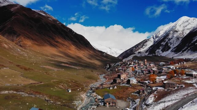 drone view of winding river cutting through valley toward scattered houses and towers, golden slopes meeting snowline, atmospheric light and expansive sky create sense of journey and remote mountain
