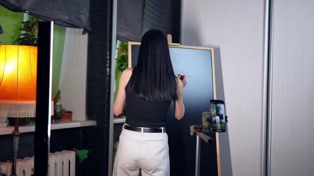 Young adult woman standing in a room and writing on the blackboard shooting the process on mobile. Back view of Caucasian female tutor preparing for the online lecture. Distant education concept