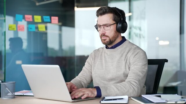 Focused businessman wearing wireless headphones typing on laptop while sitting at desk in office. Concentrated professional working on computer as IT programmer, writing code and developing software.